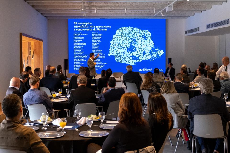 Auditório com dezenas de pessoas assistindo a apresentação; tela azul exibe mapa dos municípios do Paraná.