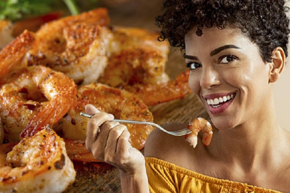 Fotografia colorida: mulher negra sorridente, blusa mostarda, leva camarão ao garfo diante de camarões grelhados temperados.