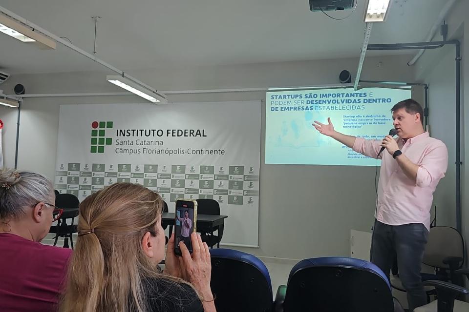 Palestra em sala do Instituto Federal de Santa Catarina – Câmpus Florianópolis-Continente. Um homem de camisa rosa clara e calça escura fala ao microfone e gesticula diante de um slide com o título “Startups são importantes e podem ser desenvolvidas dentro de empresas estabelecidas”. Duas mulheres estão sentadas na plateia; uma delas filma a apresentação com o celular.