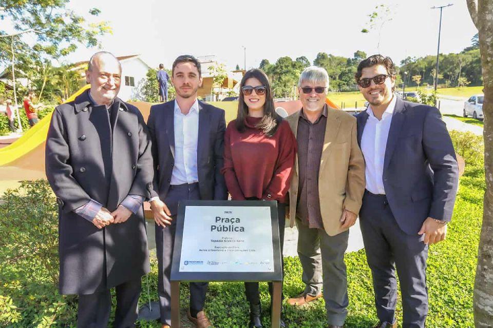 Cinco pessoas posam sorrindo em frente a uma placa de inauguração em uma praça ensolarada.