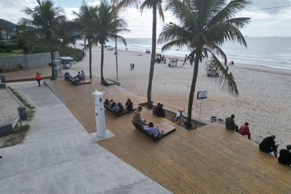 Deck de madeira à beira-mar com bancos ocupados por pessoas sentadas sob coqueiros, em frente à praia com céu parcialmente nublado.