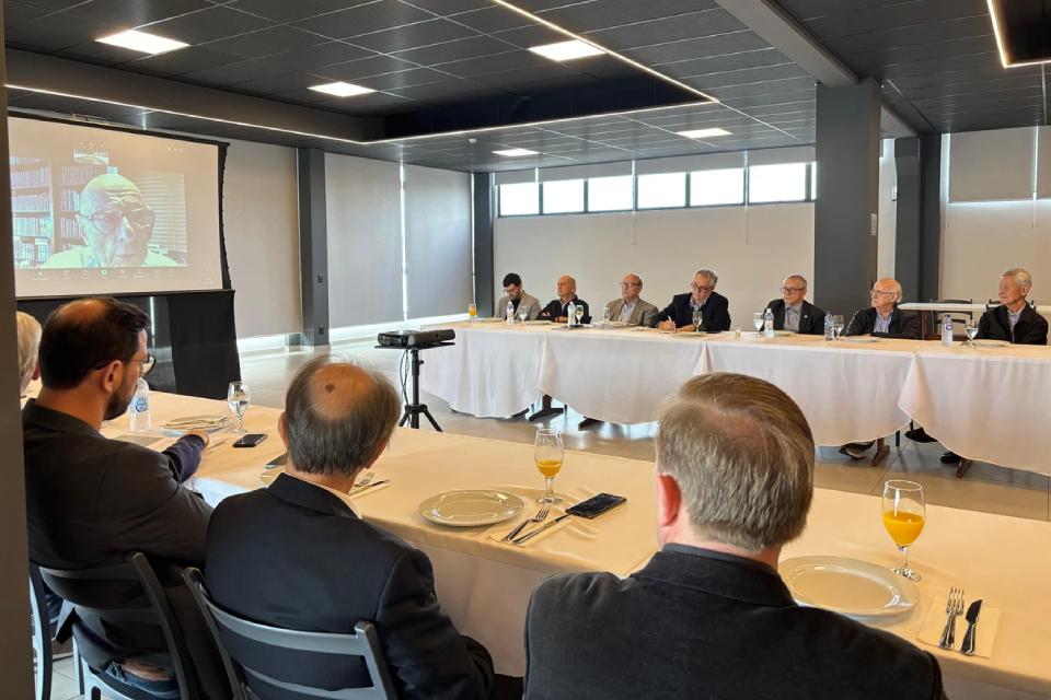 Em sala moderna, doze homens em traje social sentam-se à mesa branca com pratos e suco de laranja; participante remoto aparece projetado em telão.