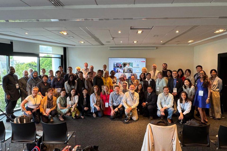 Fotografia de grupo com cerca de quarenta pessoas em sala, sorrindo diante de tela com conferência virtual.