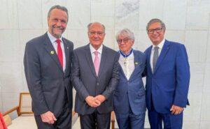 Quatro homens de terno escuro sorriem em pé, em frente a uma parede clara, em ambiente interno.