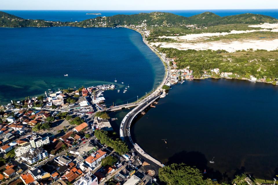 Fotografia aérea de ponte curva ligando lagoa ao mar azul, com casas à beira e morros verdes ao fundo.