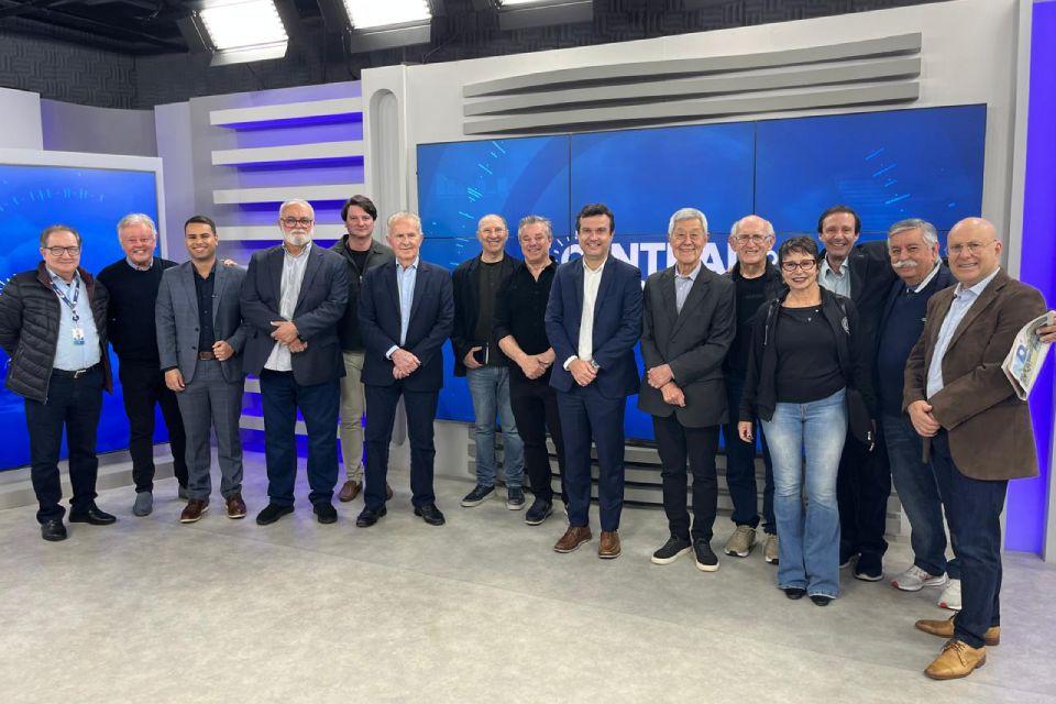 Fotografia de estúdio de TV com quinze pessoas alinhadas, formais e casuais, diante de painéis azuis.