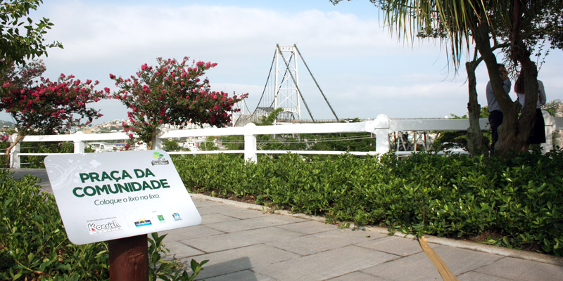 Placa da “Praça da Comunidade” em calçada cercada por vegetação e árvores floridas, com a Ponte Hercílio Luz ao fundo.