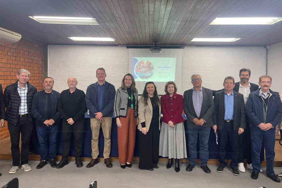 Grupo de onze pessoas em pé, lado a lado, posando para foto em uma sala de reuniões. Ao fundo há uma tela de projeção com o logotipo do Banco de Alimentos de Santa Catarina e texto de apresentação. As pessoas estão sorrindo e usam roupas formais e sociais.