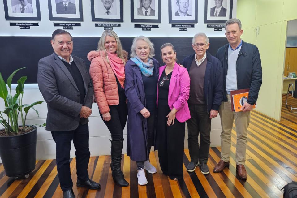 Seis pessoas em pé, lado a lado, posam sorridentes em um ambiente interno com piso de madeira listrado. Atrás deles há uma parede com retratos em preto e branco de antigos dirigentes, acompanhados de nomes e datas. Da esquerda para a direita: um homem de blazer cinza escuro, uma mulher de jaqueta rosa, uma mulher de casaco azul marinho com cachecol colorido, uma mulher de blazer rosa, um homem de suéter escuro com blazer cinza e outro homem de jaqueta preta segurando uma pasta laranja. À esquerda há um vaso preto com uma planta verde.