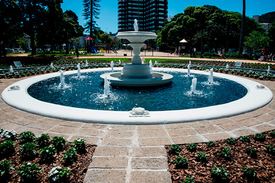 Fonte circular com jatos d’água e estrutura central branca, cercada por canteiros floridos e bancos na praça Getúlio Vargas arborizada.