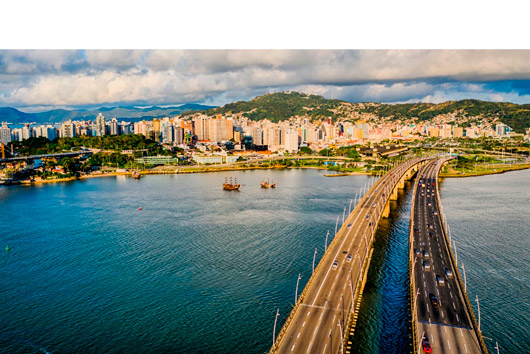 Vista aérea das duas pontes sobre o mar ligando ao centro de Florianópolis, com barcos turísticos e prédios ao fundo diante de morros verdes.