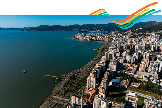 Fotografia aérea mostra a Beira-Mar Norte em Florianópolis, com prédios altos à direita e o mar azul à esquerda, cercado por morros verdes ao fundo. No topo da imagem, há um conjunto de quatro faixas curvas coloridas em laranja, amarelo, verde e azul.