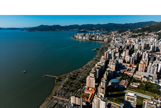 Fotografia aérea mostra a Beira-Mar Norte em Florianópolis, com prédios altos à direita e o mar azul à esquerda, cercado por morros verdes ao fundo.