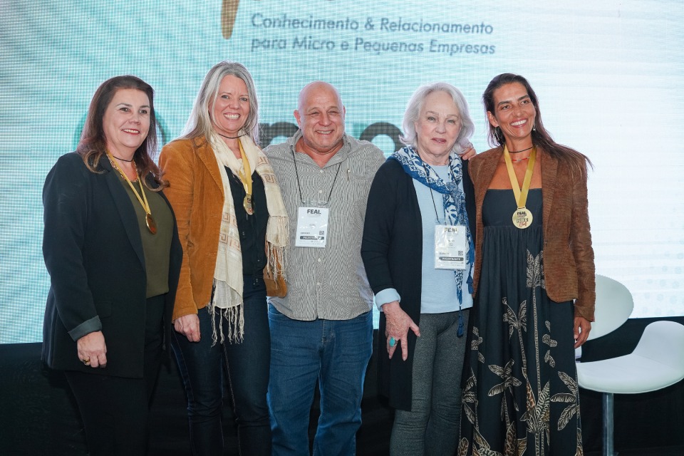 Cinco pessoas posam sorridentes lado a lado no palco, usando crachás e medalhas, diante de painel do Circuito Ampe.