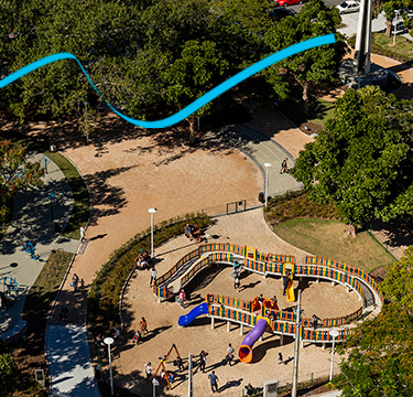 Vista aérea de uma praça arborizada com parque infantil colorido cercada por árvores de copas frondosas e verdes.