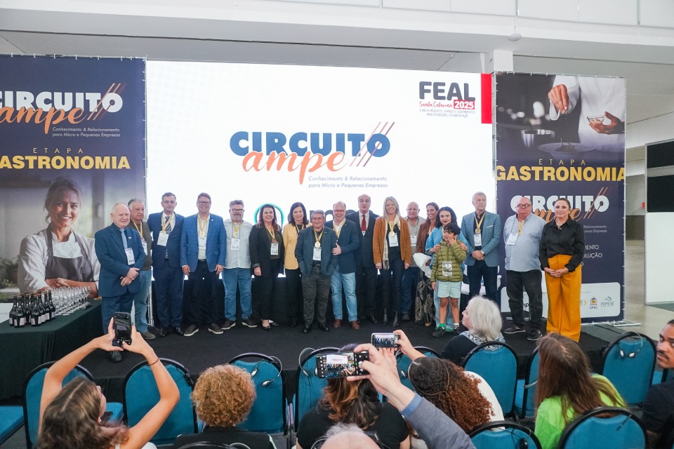 Um grande grupo de participantes reúne-se no palco em frente ao painel do Circuito Ampe, com destaque para a etapa Gastronomia.
