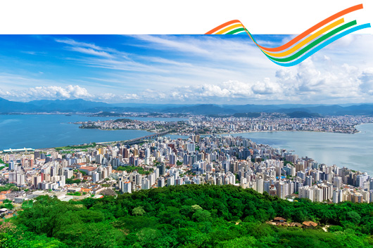 Fotografia panorâmica de Florianópolis vista do alto, mostrando o centro urbano com muitos prédios cercados pelo mar, sob um céu azul com nuvens espalhadas e vegetação verde em primeiro plano. No topo da imagem, há um conjunto de quatro faixas curvas coloridas em laranja, amarelo, verde e azul, compondo um elemento gráfico sobre o céu azul com nuvens.