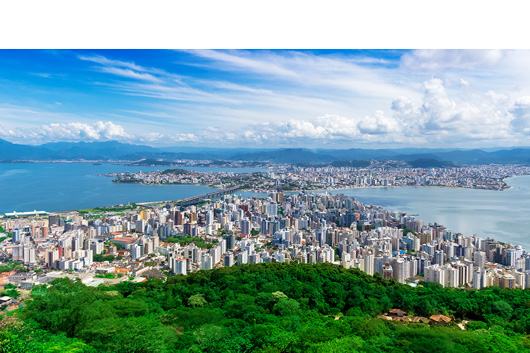 Fotografia panorâmica de Florianópolis vista do alto, mostrando o centro urbano com muitos prédios cercados pelo mar, sob um céu azul com nuvens espalhadas e vegetação verde em primeiro plano.