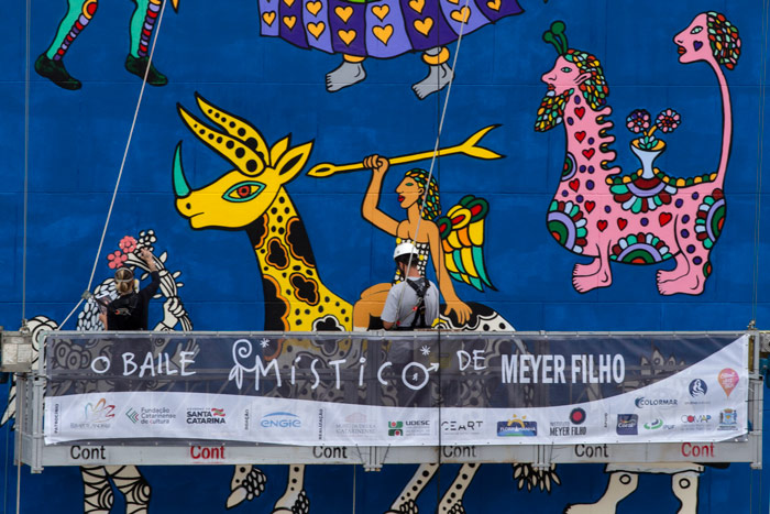 Mural colorido com fundo azul exibe figuras da obra de Meyer Filho, como uma mulher alada montando uma girafa amarela de chifres e animais estilizados em rosa e preto. Em primeiro plano, dois trabalhadores em andaime realizam pintura diante de uma faixa que diz “O Baile Místico de Meyer Filho”.