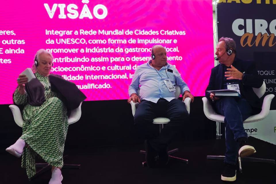 Fotografia: três palestrantes sentados em poltronas brancas, todos com headset. À esquerda, mulher de vestido verde estampado e xale; ao centro, homem de camisa azul clara; à direita, homem de blazer escuro com caderno. Fundo rosa com “VISÃO” e texto: “Integrar a Rede Mundial de Cidades Criativas da UNESCO, como forma de impulsionar e promover a indústria da gastronomia local, contribuindo assim para o desenvolvimento socioeconômico e cultural…”.