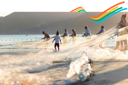 Fotografia mostra um grupo de pessoas puxando uma rede de pesca no mar raso ao amanhecer, com uma criança caminhando entre elas. No topo da imagem, há um conjunto de quatro faixas curvas coloridas em laranja, amarelo, verde e azul.