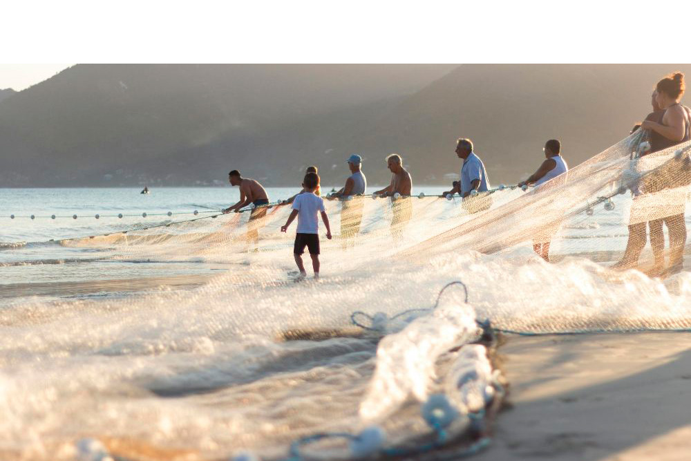 Fotografia mostra um grupo de pessoas puxando uma rede de pesca no mar raso ao amanhecer, com uma criança caminhando entre elas.