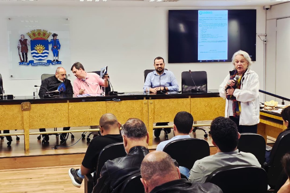 Fotografia colorida mostra uma mulher de cabelos grisalhos falando ao microfone diante de uma plateia sentada, em uma sala de reuniões oficial. Atrás dela, três homens estão sentados à mesa de madeira dourada, com o brasão da cidade de Florianópolis na parede e uma tela exibindo um documento digital.