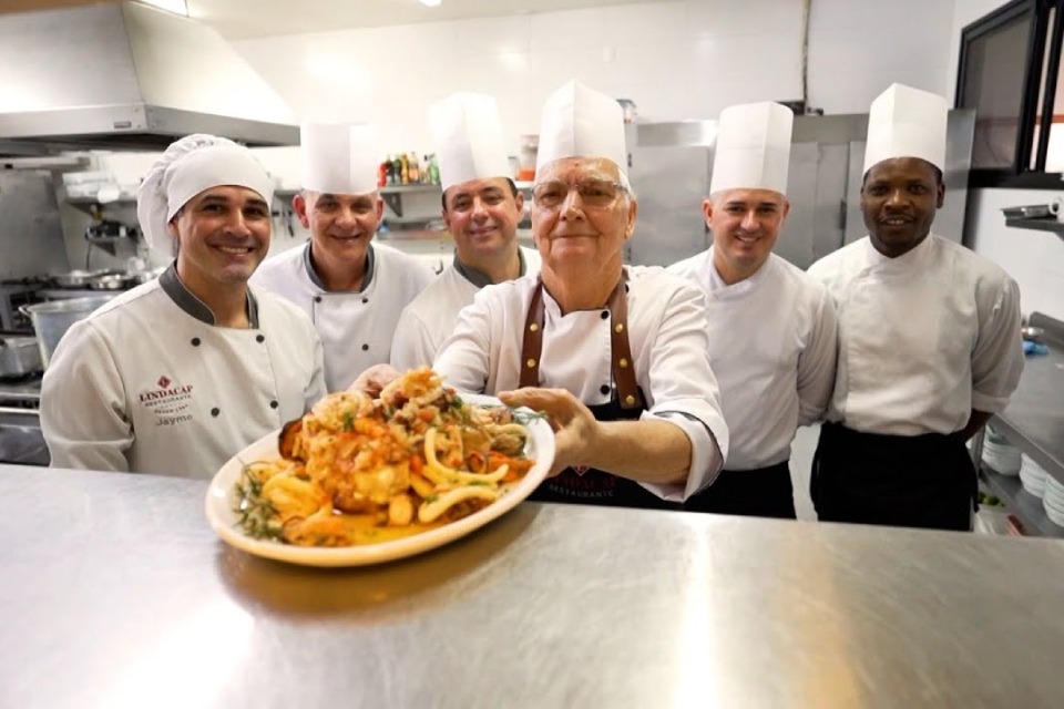 Seis chefs de cozinha sorridentes posam em uma cozinha profissional; o chef ao centro segura um prato de frutos do mar colorido em destaque.