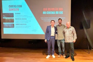 Três homens posam sorridentes diante de uma tela de projeção com o cartaz do evento “Curtas com Contexto”.