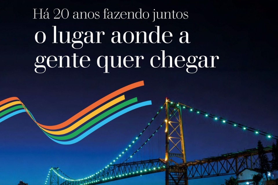 Fotografia noturna da ponte Hercílio Luz iluminada, com o texto “Há vinte anos fazendo juntos o lugar aonde a gente quer chegar” sobreposto e uma faixa colorida ondulada em tons de laranja, verde e azul.