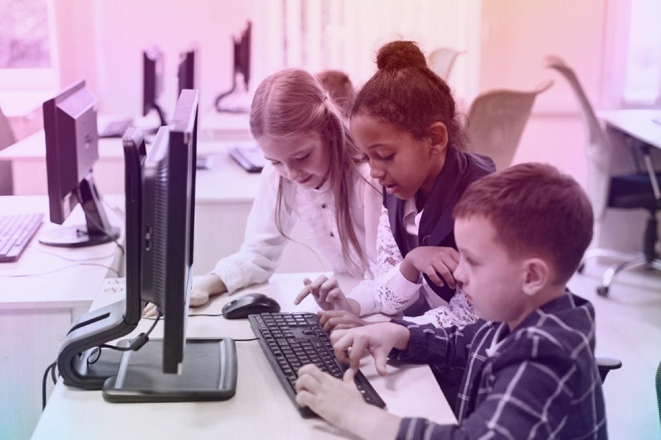 Três crianças sentadas em frente a um computador colaboram em uma atividade, concentradas e sorridentes, em uma sala de aula moderna. ”. 