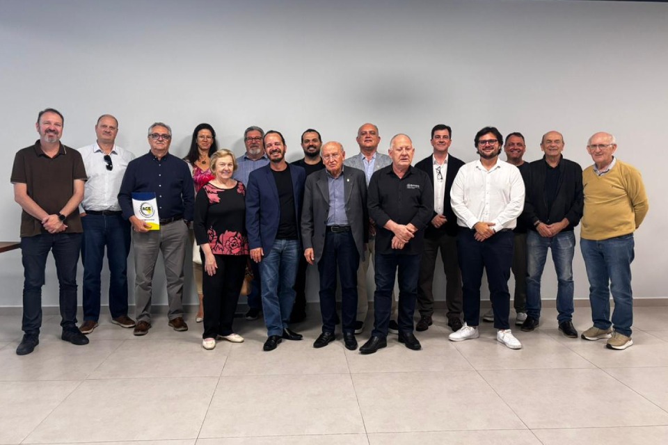 Fotografia de grupo com cerca de quinze pessoas alinhadas diante de parede clara, todas sorrindo, em ambiente interno iluminado.