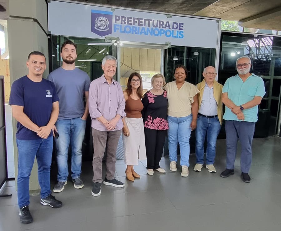 Homem de camiseta preta e crachá verde fala ao microfone diante de tela que exibe “Florianópolis 2025”, “Fotografia de oito adultos alinhados sorrindo diante da entrada da Prefeitura de Florianópolis, em ambiente interno iluminado.