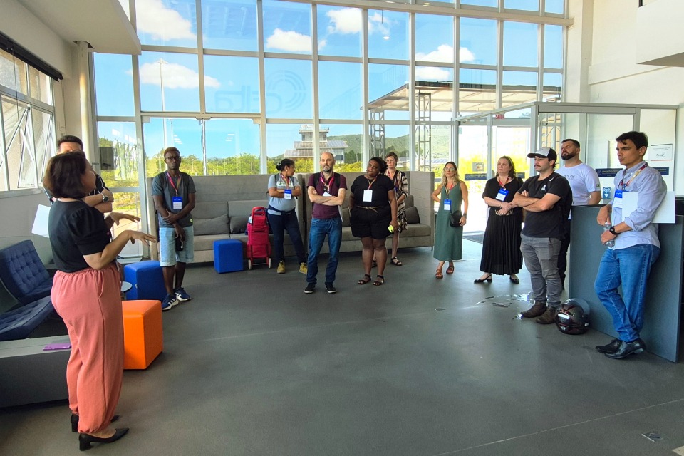 Fotografia em ambiente corporativo iluminado, onde uma mulher fala para onze pessoas em círculo, todas usando crachás, ouvindo atentamente em um espaço moderno com janelas amplas.