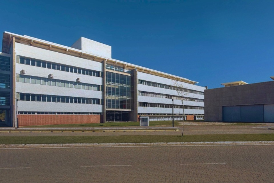 Fotografia externa de um edifício moderno de vários andares, com fachada branca e janelas horizontais, céu azul ao fundo e área gramada e pavimentada em frente ao prédio.
