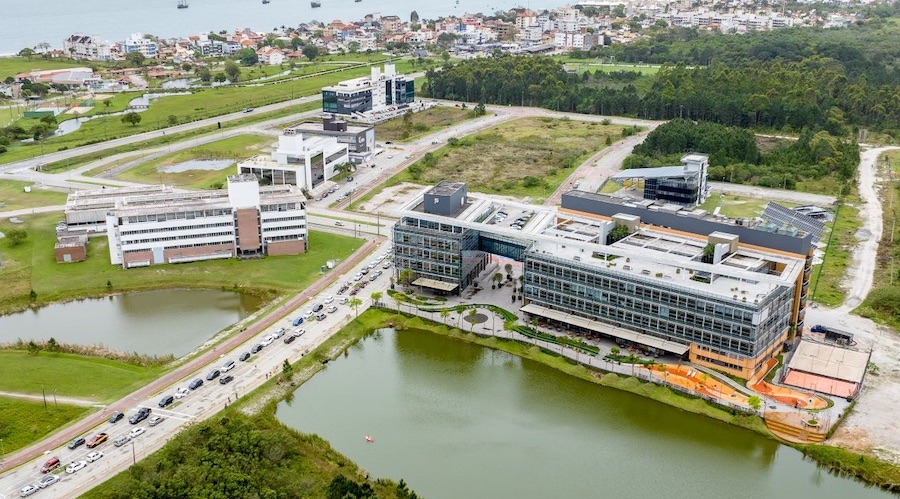 Fotografia aérea de parque tecnológico com prédios modernos à beira de um lago, cercados por áreas verdes, vias internas e bairro urbano ao fundo.