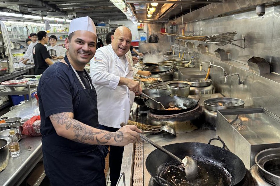 Fotografia em cozinha industrial: cozinheiro de touca e avental sorri com concha no wok, ao lado de chef de branco rindo; panelas fumegantes e equipe ao fundo.