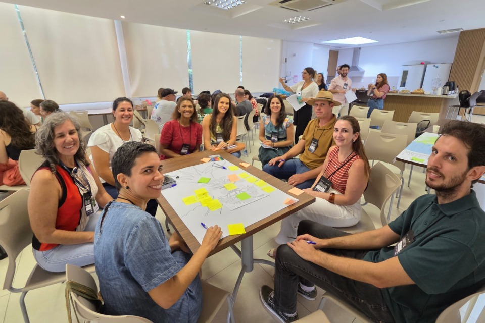 Fotografia de oficina colaborativa com nove participantes sorrindo ao redor de uma mesa com post-its em sala de evento.