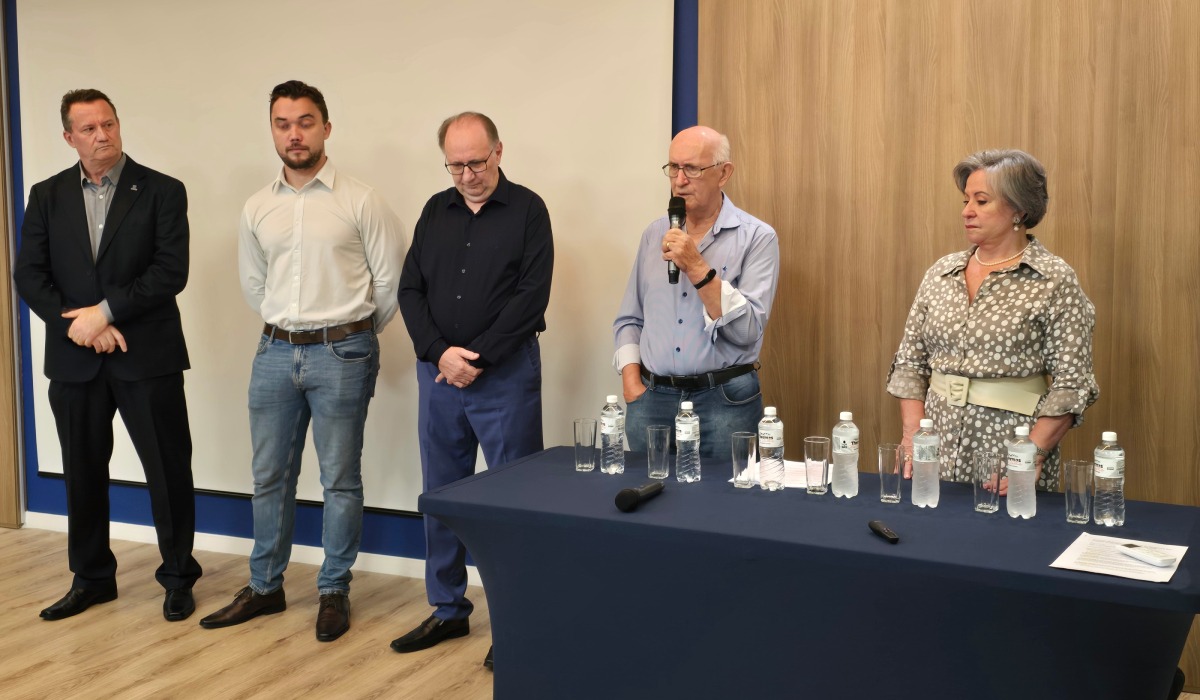 Fotografia de evento em sala: cinco pessoas em pé atrás de uma mesa com garrafas e copos, enquanto um homem idoso fala ao microfone.