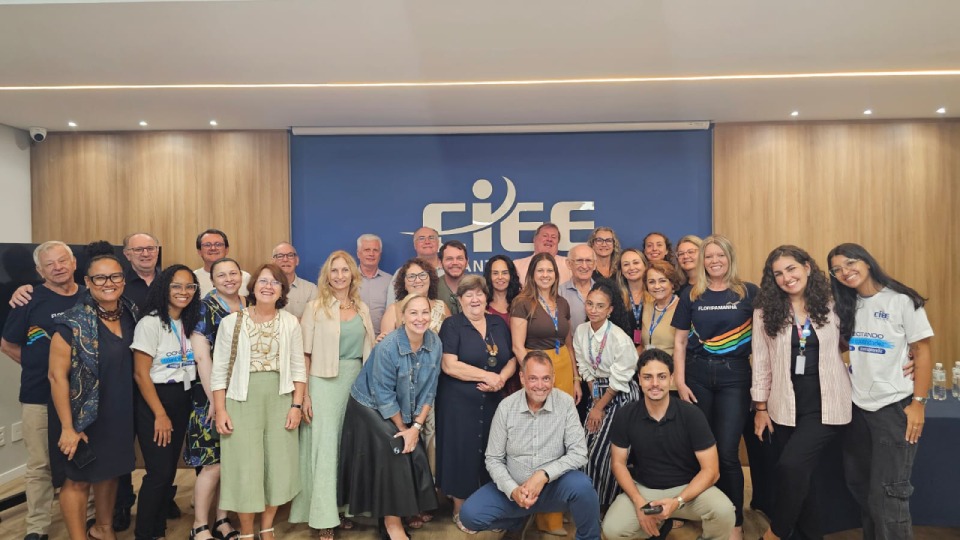 Fotografia de grupo com cerca de trinta pessoas sorrindo em uma sala de eventos, posadas diante de um painel azul com o logotipo do CIEE. O grupo reúne homens e mulheres de idades variadas, com roupas casuais e crachás, distribuídos em pé e agachados na primeira fila.