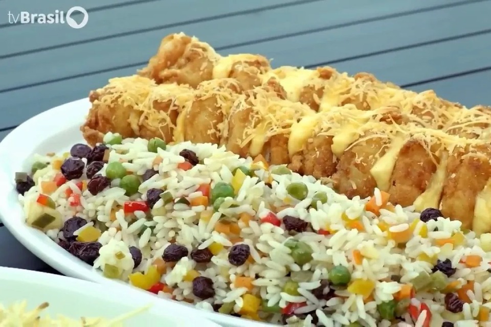 Fotografia de travessa branca com arroz colorido, ervilhas e uvas-passas ao lado de frango dourado com molho e queijo ralado; no canto superior esquerdo, logotipo “tv Brasil”.