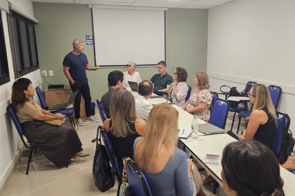 Fotografia de reunião em sala: homem em pé fala a um grupo sentado em volta de mesas, enquanto uma mulher registra a cena com o celular.