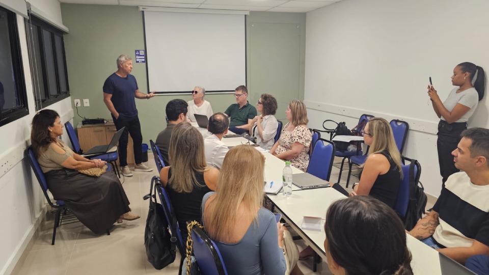 Fotografia de reunião em sala: homem em pé fala a um grupo sentado em volta de mesas, enquanto uma mulher registra a cena com o celular.