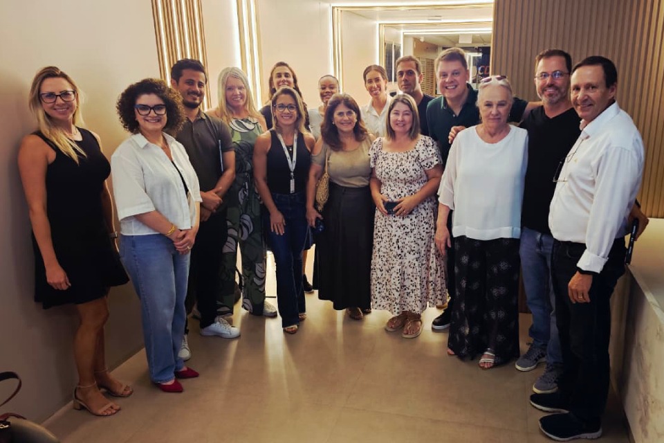 Fotografia de quinze adultos em pé, lado a lado, posando sorridentes em um corredor interno bem iluminado.k