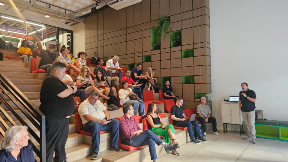 Fotografia de palestra em auditório moderno, com dezenas de pessoas na arquibancada e um homem falando ao microfone.