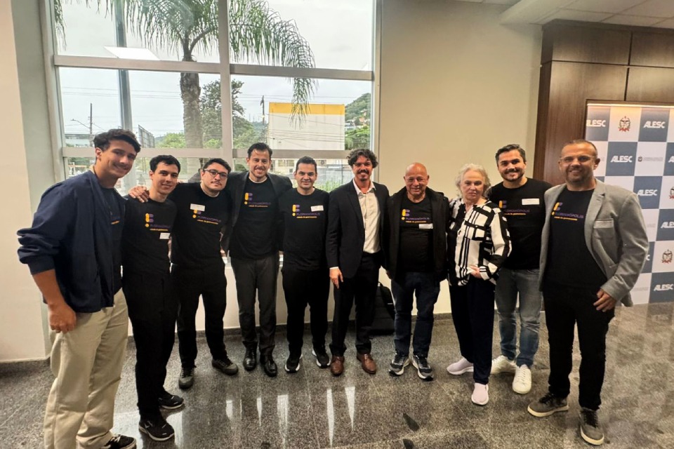 Fotografia de grupo com nove homens e uma mulher em ambiente interno, diante de janelas e painel da Alesc.