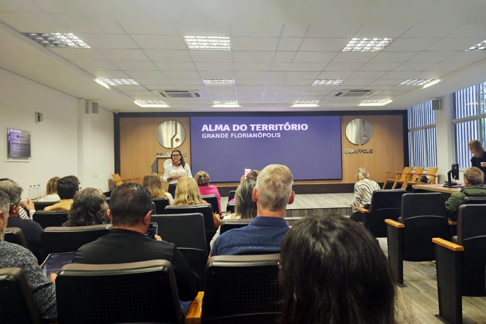 Fotografia de auditório com público acompanhando apresentação no palco, diante de telão com o texto “Alma do Território Grande Florianópolis”