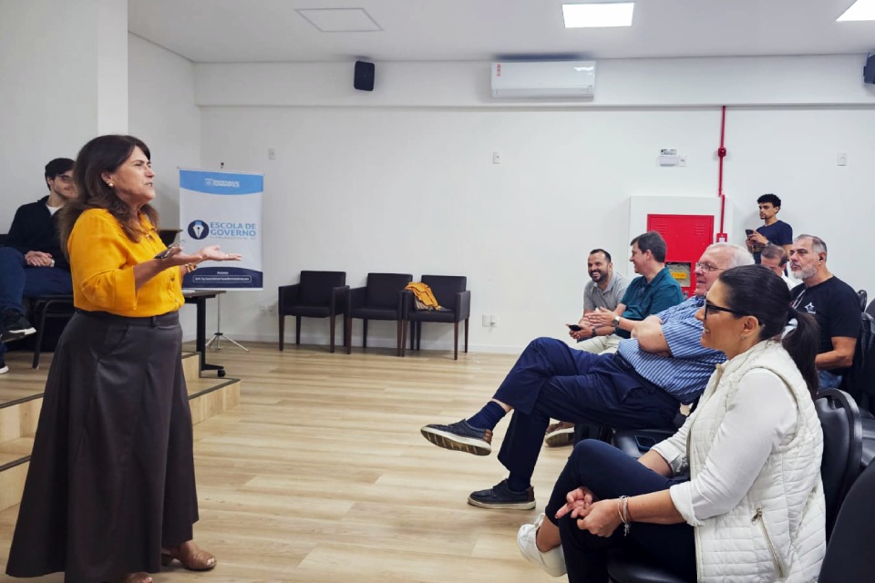 Fotografia de uma mulher em pé falando a um grupo sentado em sala de reunião da Escola de Governo.