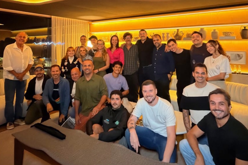 Fotografia de grupo com vinte e três pessoas sorrindo em uma sala aconchegante, com sofá branco e iluminação amarela. 
