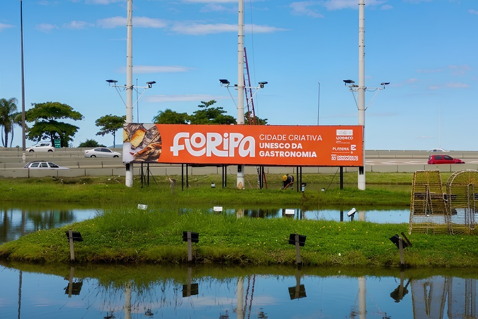 "Fotografia de lago com reflexo do céu e de um gramado central, onde há um painel laranja com o texto “FLORIPA CIDADE CRIATIVA UNESCO DA GASTRONOMIA”